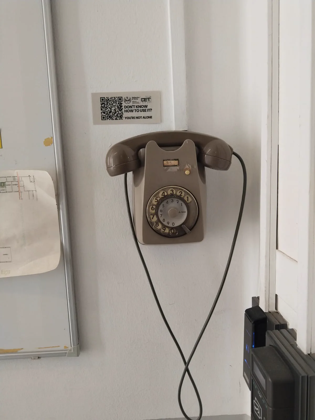 Politecnico'da kapı yanında duran eski tip çevirmeli telefon ve QR bilgilendirme etiketi